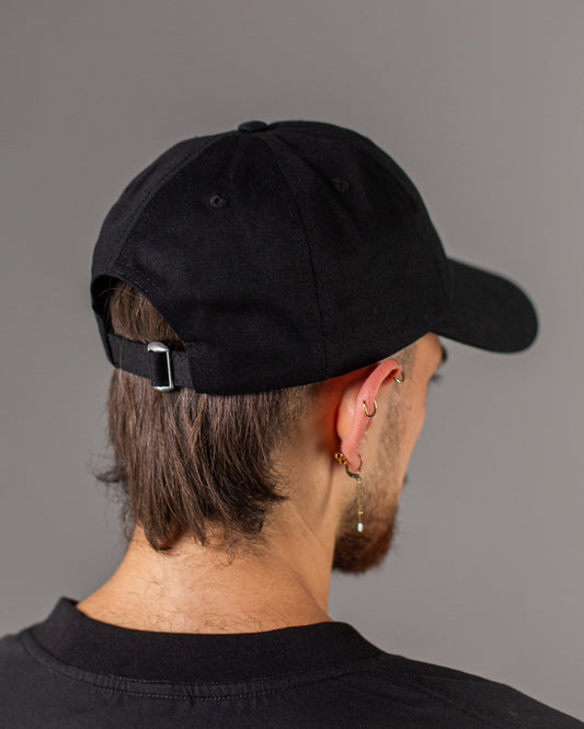 Rückansicht der getragenen schwarzen Cap mit geschlossenem Stoffband und silberfarbener Metallschnalle zur Größenverstellung, Haare ragen unter der Cap hervor.