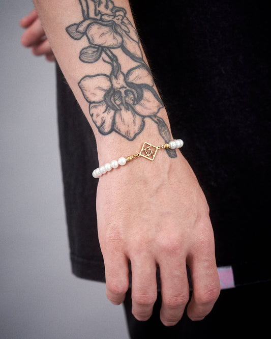 Nahaufnahme eines Handgelenks mit großflächigem Blumentattoo, an dem ein elastisches Armband mit weißen Perlen und einem goldfarbenen Logo-Element von LOKEY getragen wird.