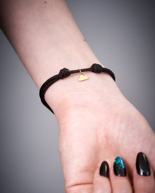 Nahaufnahme eines schwarzen Armbands mit kleinem goldfarbenem Wolkenanhänger am Handgelenk einer Person. Das Band ist doppelt geknotet und minimalistisch gestaltet.