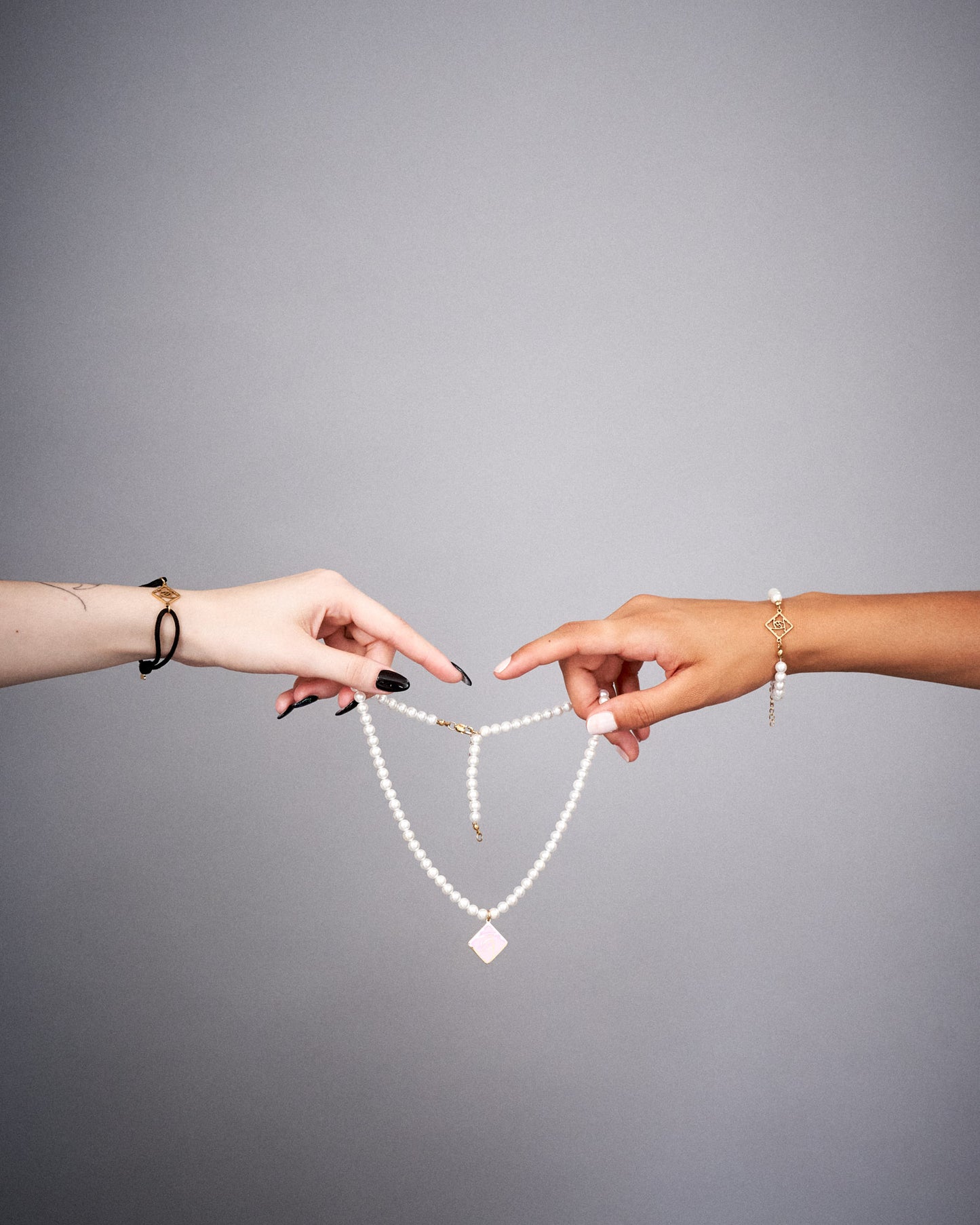 Eine hellhäutige und eine dunkelhäutige Hand halten die weiße Perlenkette in der Luft. Der Anhänger in Rautenform mit rosa Rosensymbol hängt mittig. Beide Handgelenke tragen Schmuck, darunter ein passendes Perlenarmband.