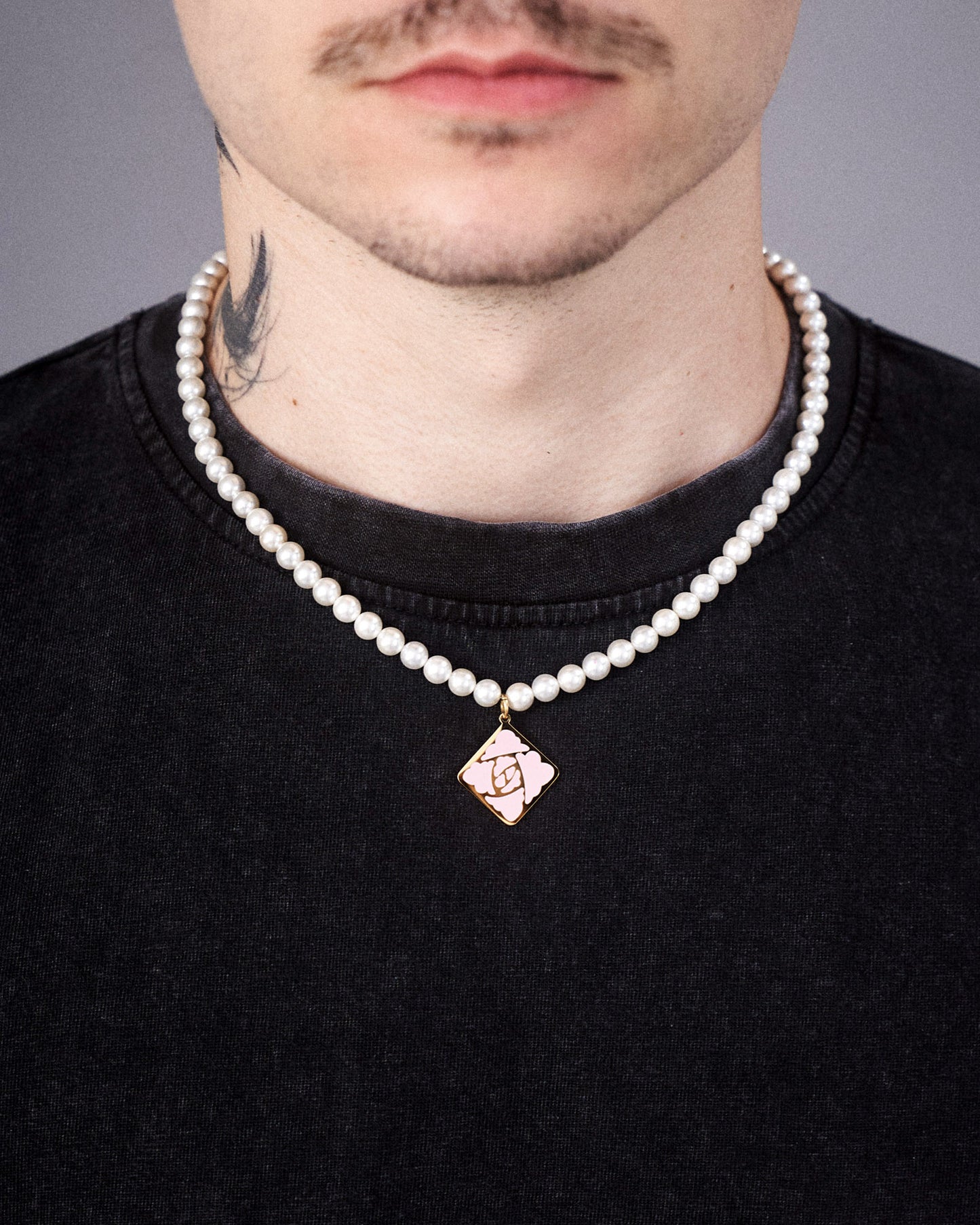 Die weiße Perlenkette mit mittigem Rosensymbol-Anhänger in Rautenform wird um den Hals getragen. Der goldene Anhänger hebt sich deutlich vom dunklen T-Shirt ab. Sichtbar ist auch ein Teil eines Tattoos am Hals.