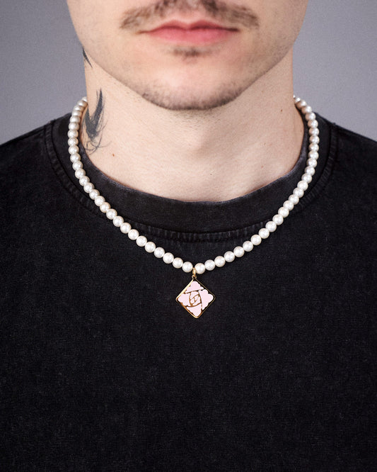 Die weiße Perlenkette mit mittigem Rosensymbol-Anhänger in Rautenform wird um den Hals getragen. Der goldene Anhänger hebt sich deutlich vom dunklen T-Shirt ab. Sichtbar ist auch ein Teil eines Tattoos am Hals.