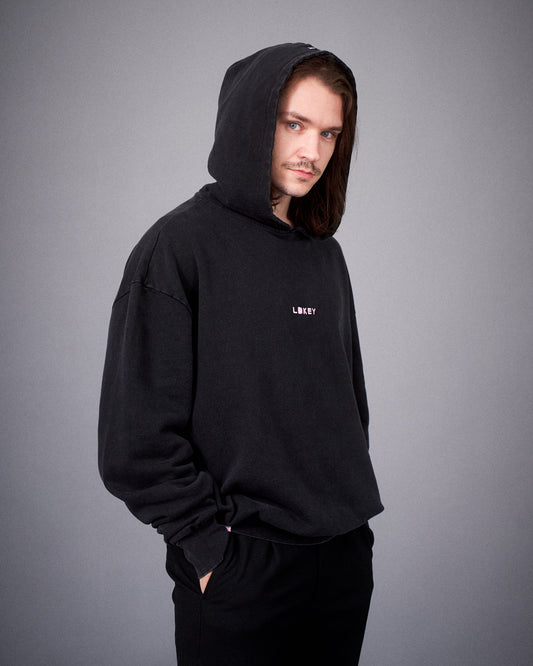 Person mit schwarzem Hoodie und aufgesetzter Kapuze blickt seitlich in die Kamera; die rosa Stickerei „LOKEY“ ist auf der Brust sichtbar.