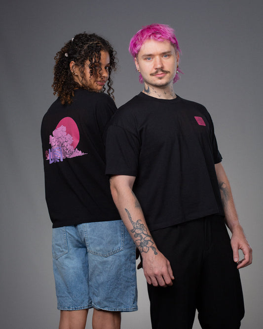 Zwei Personen stehen Rücken an Rücken und tragen das gleiche schwarze T-Shirt mit pinkem Front- und Rückendruck. Der Rückendruck zeigt eine große Wolke mit pinker Sonne und lilafarbenem „LOKEY“-Schriftzug, der Frontprint ein rechteckiges Blumenlogo in Pink.