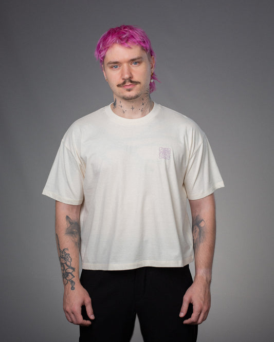 Person trägt ein cremefarbenes T-Shirt mit lockerem Schnitt. Auf der linken Brust befindet sich ein kleiner, quadratischer Print in helllila mit floralem Linienmuster.