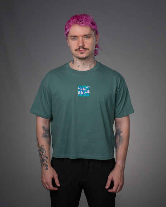 Model trägt ein salbeigrünes T-Shirt mit lockerem Schnitt. Auf der Brust ist mittig ein quadratischer Patch mit grafischer Stickerei in Blau, Weiß und Türkis angebracht. Das Motiv besteht aus abstrakten Linienformen und Wolkenelementen.