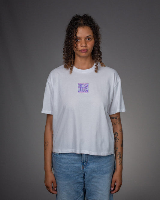 Weißes T-Shirt mit lilafarbener Stickerei auf der Brust, getragen von einer Person mit lockigen Haaren und Jeans. Frontalansicht vor grauem Hintergrund.