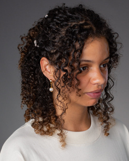 Seitenansicht eines goldfarbenen Ohrrings mit weißer Perle am Ohr einer Person mit dunklen Locken und hellem Oberteil.