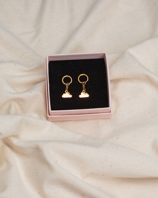 Goldene Hängeohrringe mit kleinen Wolkenanhängern, präsentiert in einer rosa Schmuckbox mit schwarzem Inlay auf naturfarbenem Stoff.