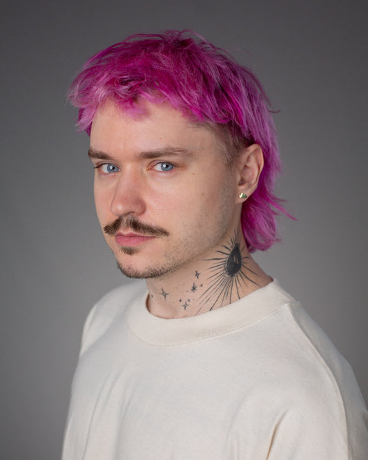 Nahaufnahme eines männlichen Models mit pinkem Haar und naturweißem Shirt. Im linken Ohr trägt er einen kleinen, goldfarbenen Ohrstecker in Wolkenform.