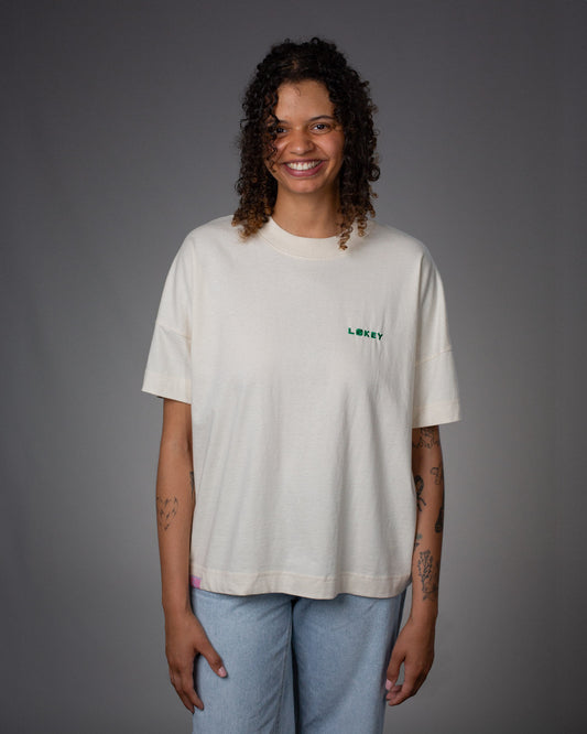 Das naturweiße T-Shirt zeigt links auf der Brust einen gestickten „LÓKEY“-Schriftzug in Grün mit dekorativem Logo im „O“. Das Model trägt das Shirt locker über hellblauer Jeans.