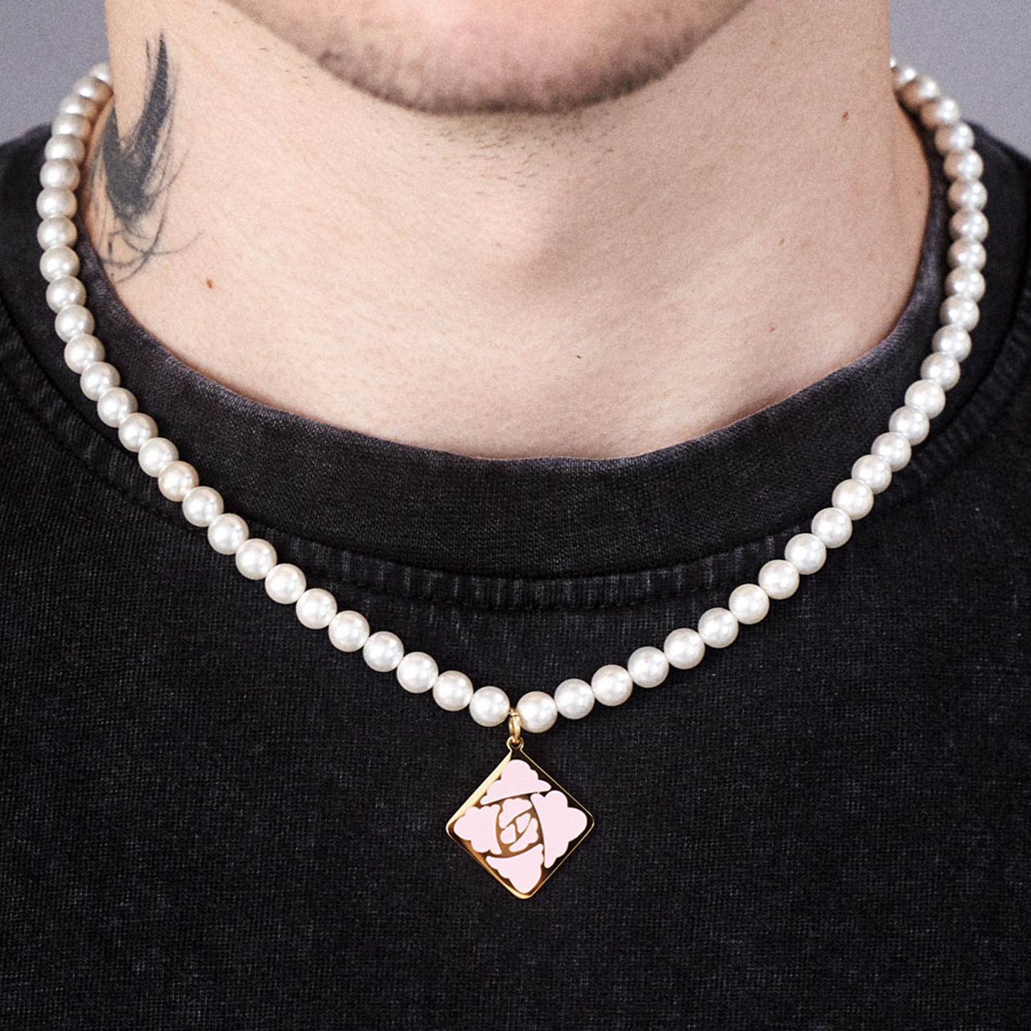 Die Kette besteht aus gleichmäßig gereihten weißen Perlen und trägt mittig einen goldenen Anhänger in Rautenform. Der Anhänger zeigt das rosa LOKEY-Rosensymbol auf schwarzem Hintergrund. Die Kette liegt über einem schwarzen T-Shirt.