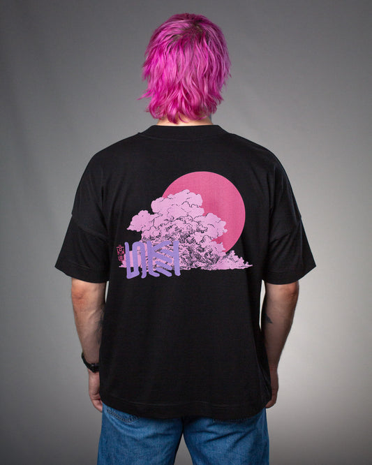 Schwarzes T-Shirt mit auffälligem Rückendruck: Eine große Baumwolke in rosa-violettem Farbverlauf vor einer pinken Kreisfläche, kombiniert mit violetter Schrift in japanischem Stil und rotem Siegelzeichen. Getragen von einer Person mit pink gefärbten Haaren und blauer Jeans.