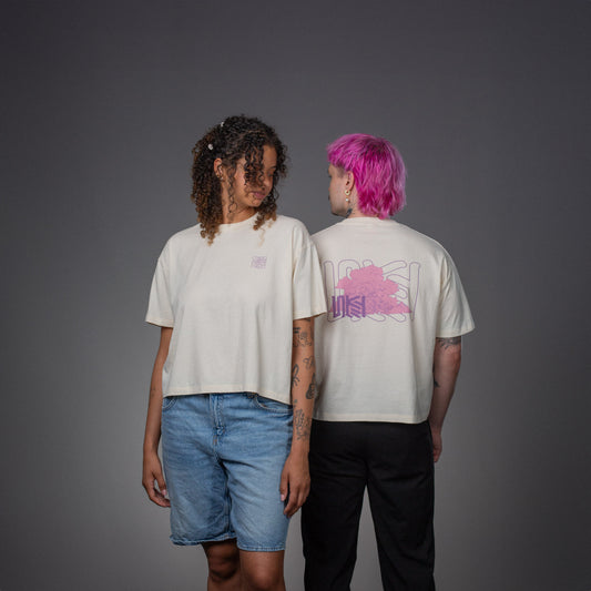 Zwei Personen tragen das cremefarbene LOKEY T-Shirt mit lockerem Schnitt. Die vordere Person zeigt den Brustprint mit lilafarbenem, quadratischem Linienmotiv. Die hintere Person steht mit dem Rücken zur Kamera, sichtbar ist der großflächige Rückendruck mit rosa Wolkenillustration und violettem „LOKEY“-Schriftzug.