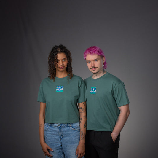 Zwei Models tragen das salbeigrüne T-Shirt in unterschiedlichen Schnitten. Beide T-Shirts zeigen mittig auf der Brust das quadratische Stickmotiv in Blau, Weiß und Türkis. Die Passform unterscheidet sich leicht: ein reguläres Unisex-Modell und ein leicht gecropptes Modell.
