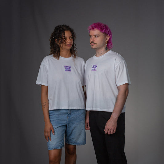  Zwei Personen tragen weiße T-Shirts mit identischer lilafarbener Stickerei auf Brusthöhe. Die linke Person trägt Jeansshorts, die rechte schwarze Hose. Beide stehen frontal im neutralen Studio-Setting.
