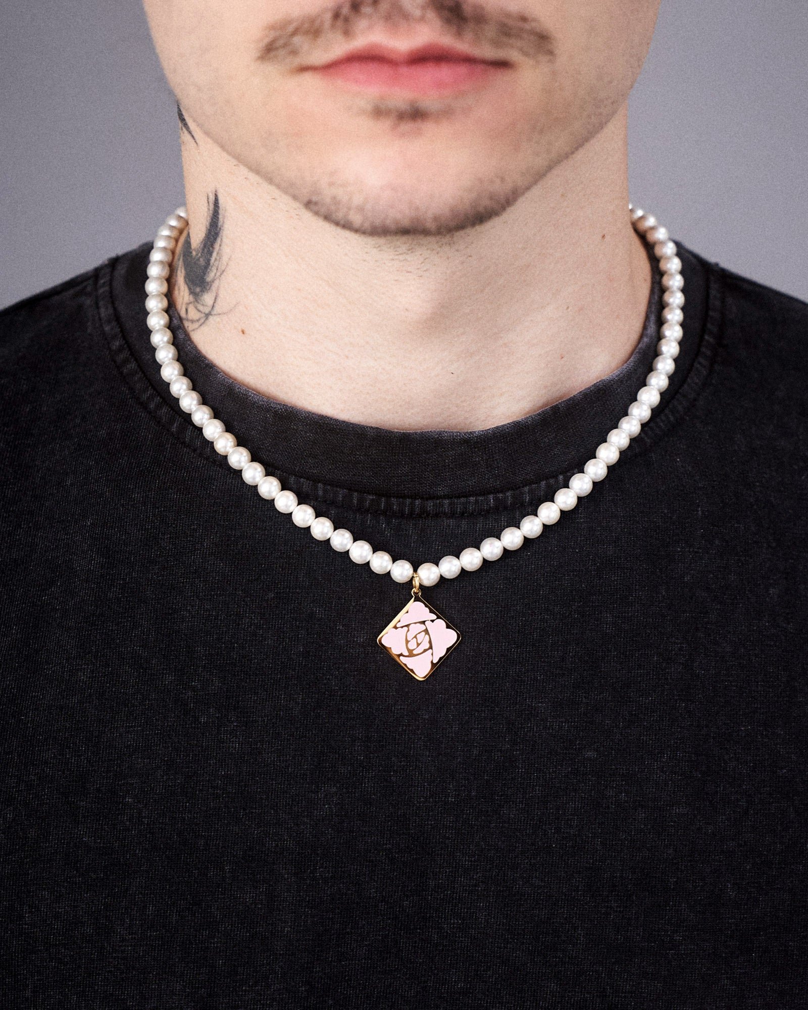 Die weiße Perlenkette mit mittigem Rosensymbol-Anhänger in Rautenform wird um den Hals getragen. Der goldene Anhänger hebt sich deutlich vom dunklen T-Shirt ab. Sichtbar ist auch ein Teil eines Tattoos am Hals.