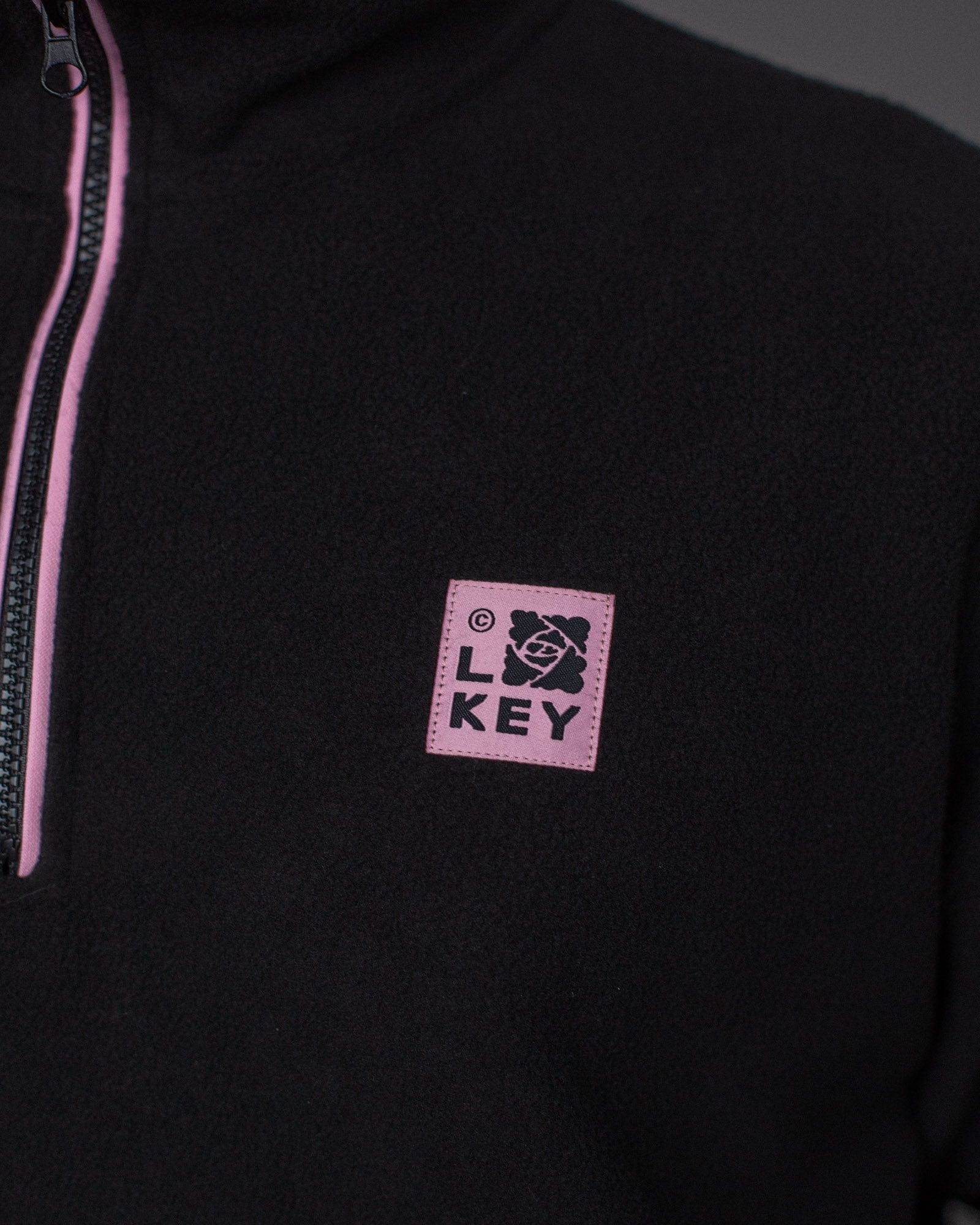 Nahaufnahme des rosa „LOKEY“-Logopatches auf der linken Brust des schwarzen Fleece-Troyers.
