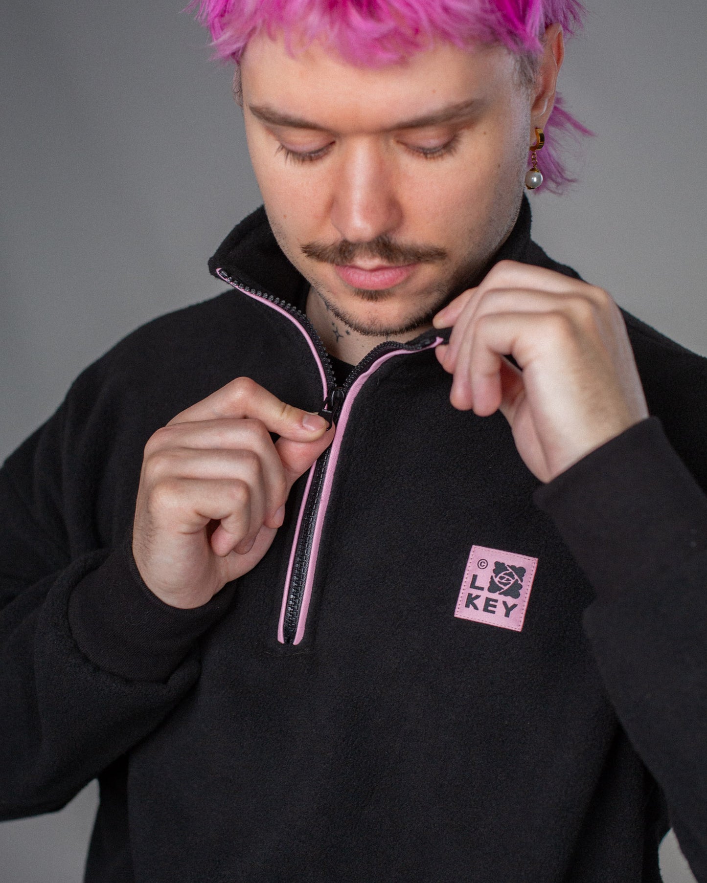 Der schwarze Fleece-Troyer wird teilweise geöffnet, der rosa unterlegte Reißverschluss mit schwarzer Naht ist sichtbar.
